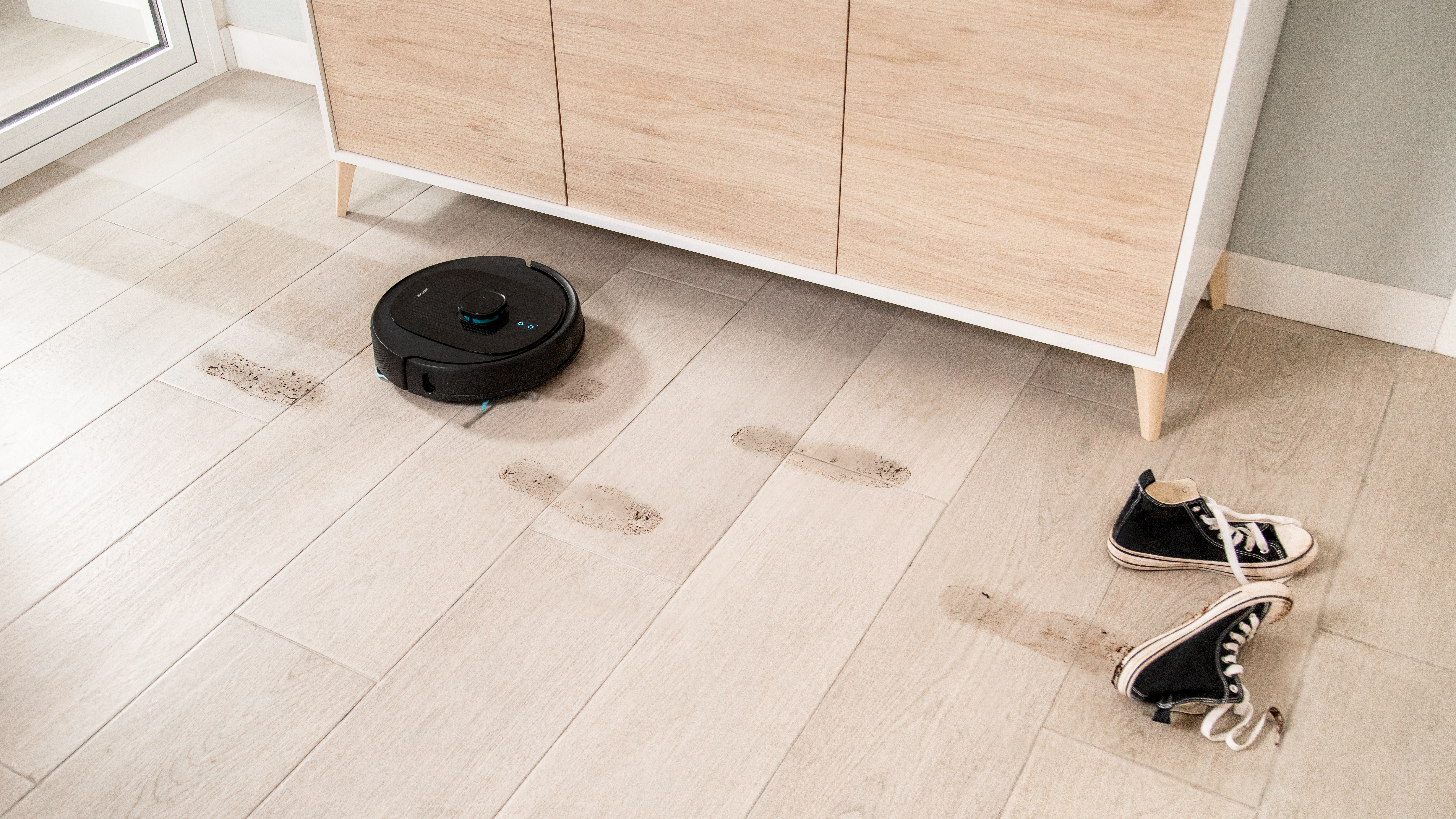 Robot aspirador, suelo sucio y zapatos. Manchas marrones y suelo de madera clara.