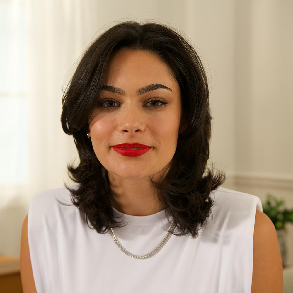 Eine Frau mit dunklen Haaren, rotem Lippenstift und weißem Oberteil lächelt.