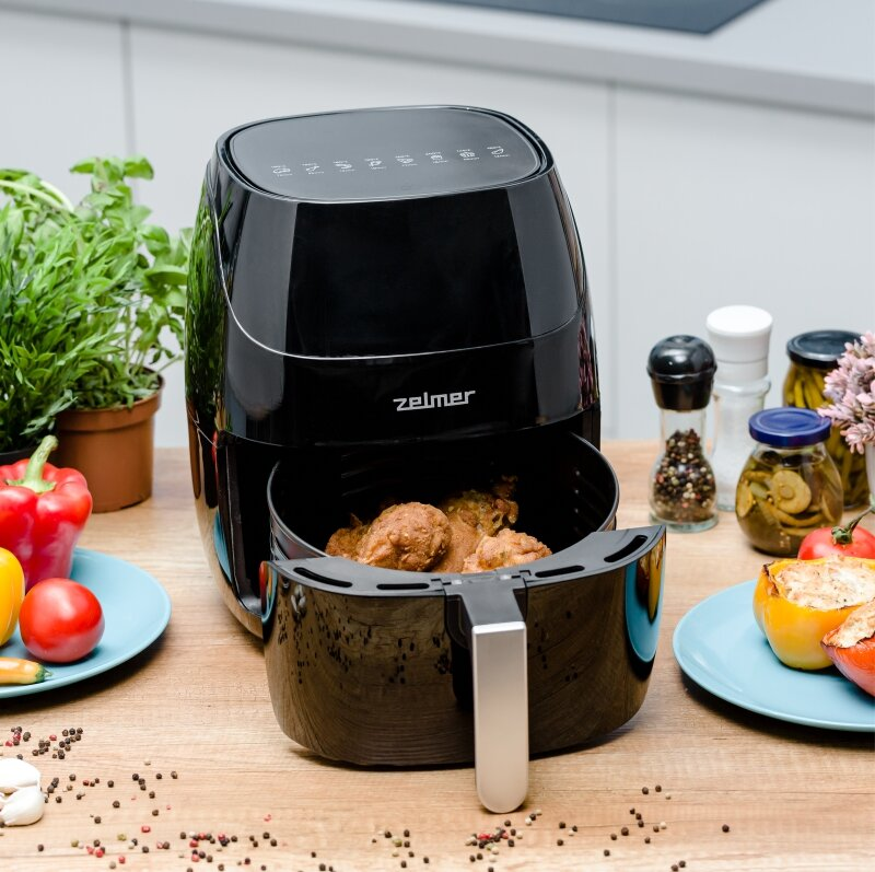 Czarna frytkownica powietrzna z jedzeniem w środku, otoczona warzywami i przyprawami.