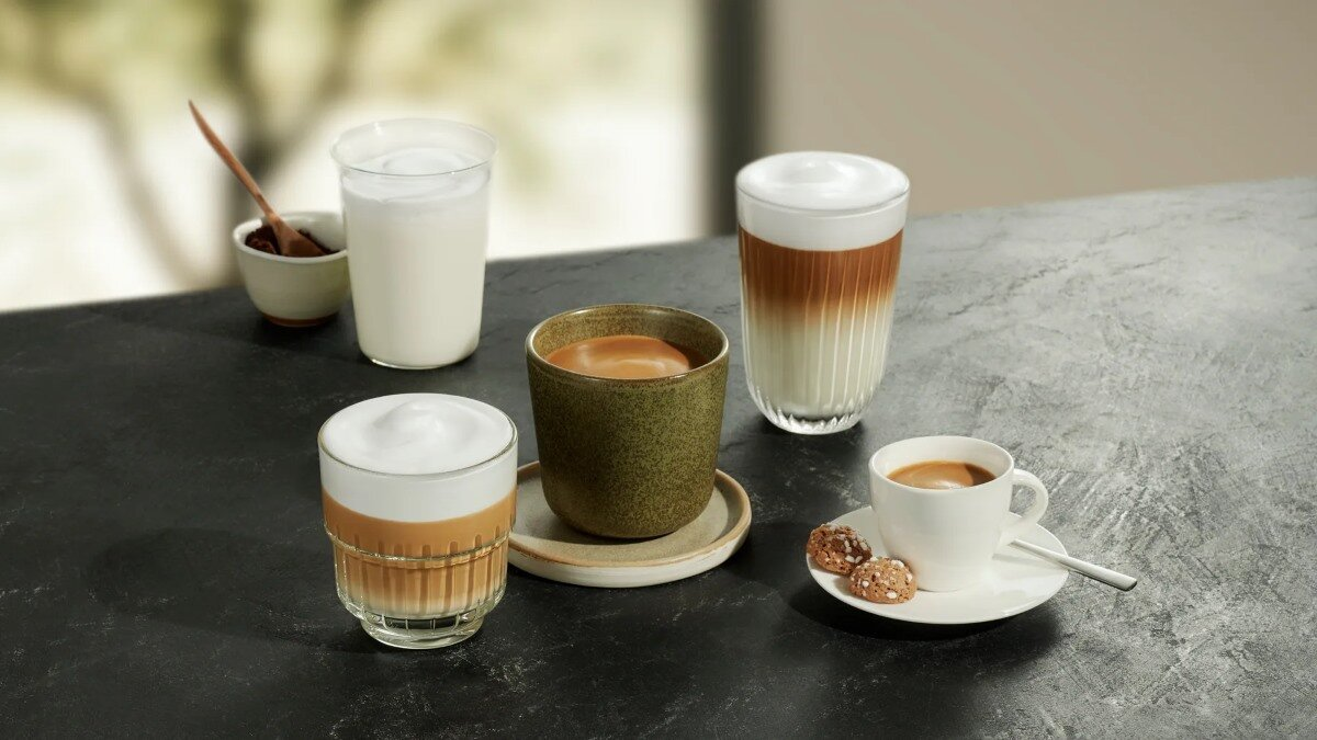 Pięć filiżanek napojów kawowych na ciemnej powierzchni. Obecna jest łyżka i ciastka.