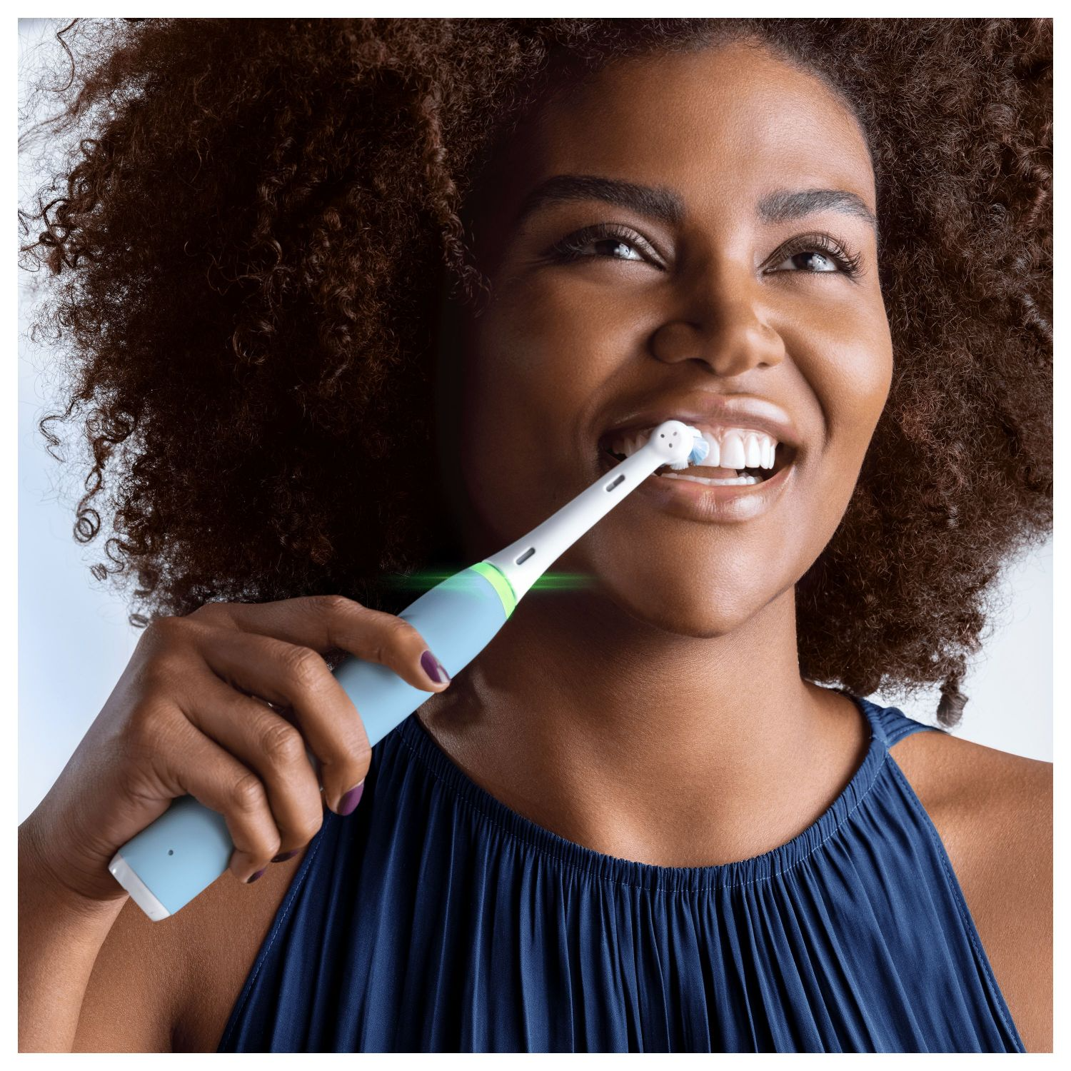 Woman brushes teeth with blue electric toothbrush. Focus on teeth, blue top, and curly hair.