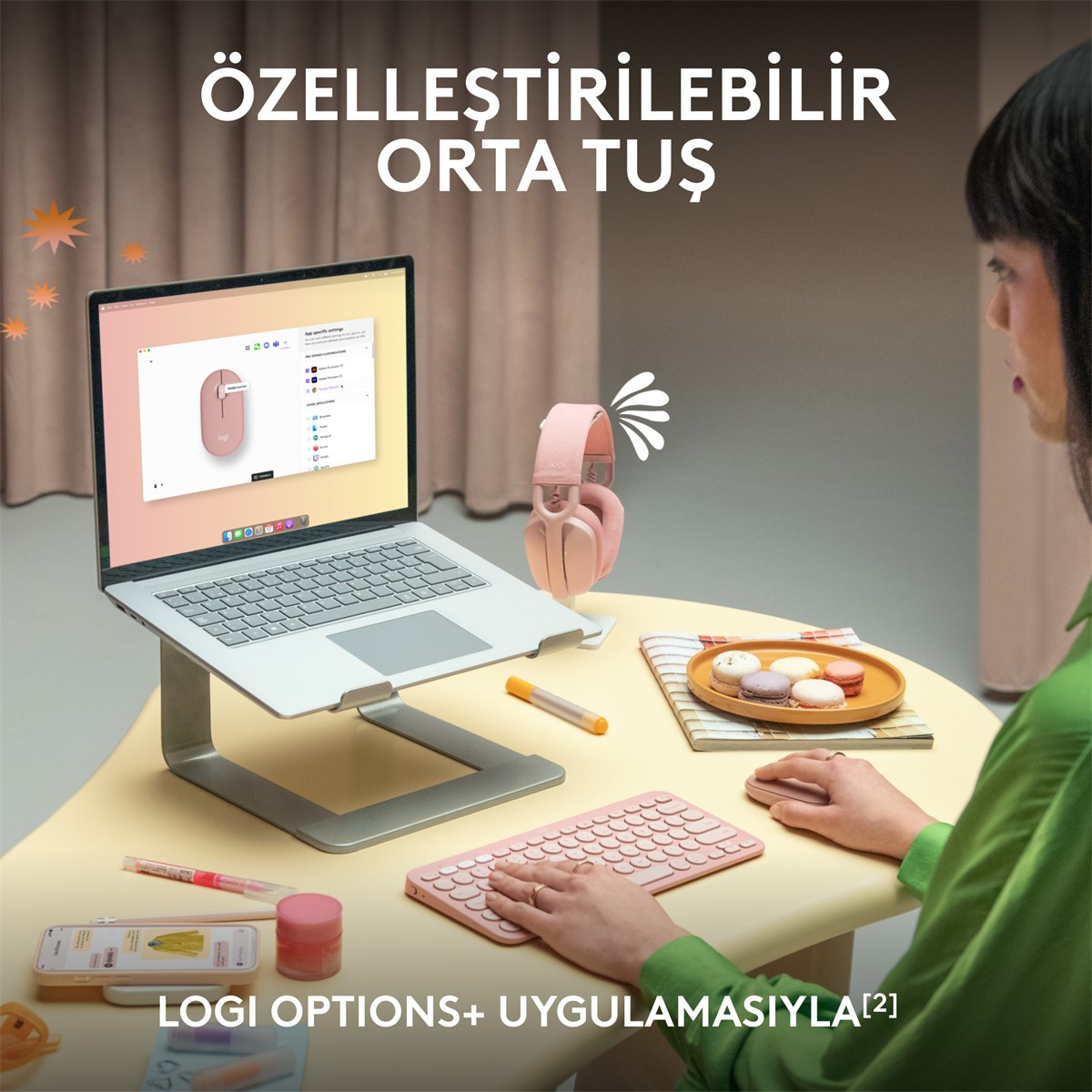 Bir kadın, bir stand üzerinde dizüstü bilgisayar kullanıyor. Pembe bir klavye, kulaklık ve telefon masada.