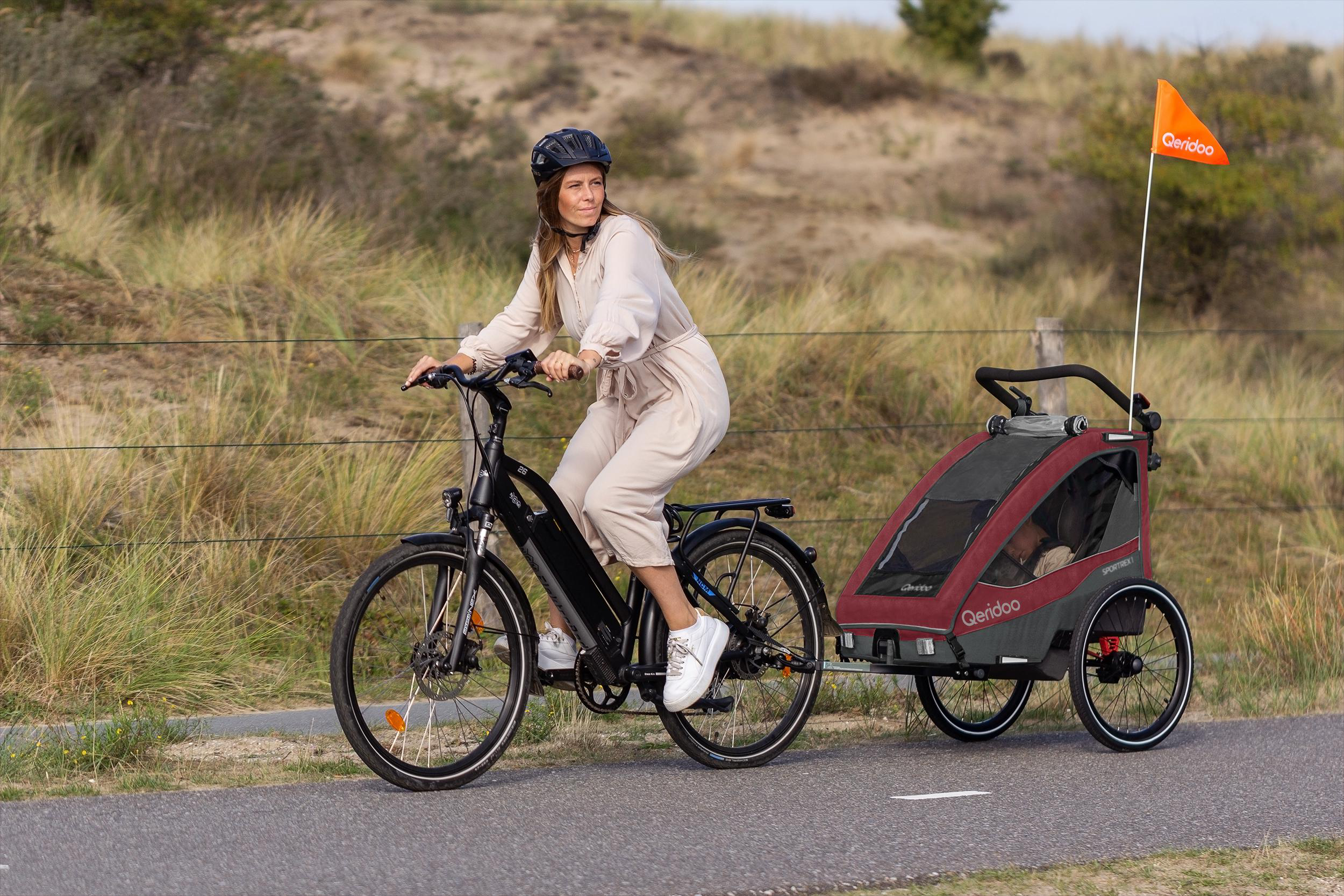 Eine Frau fährt mit einem schwarzen E-Bike, das einen Kindertrailer auf einem asphaltierten Weg am Meer zieht.