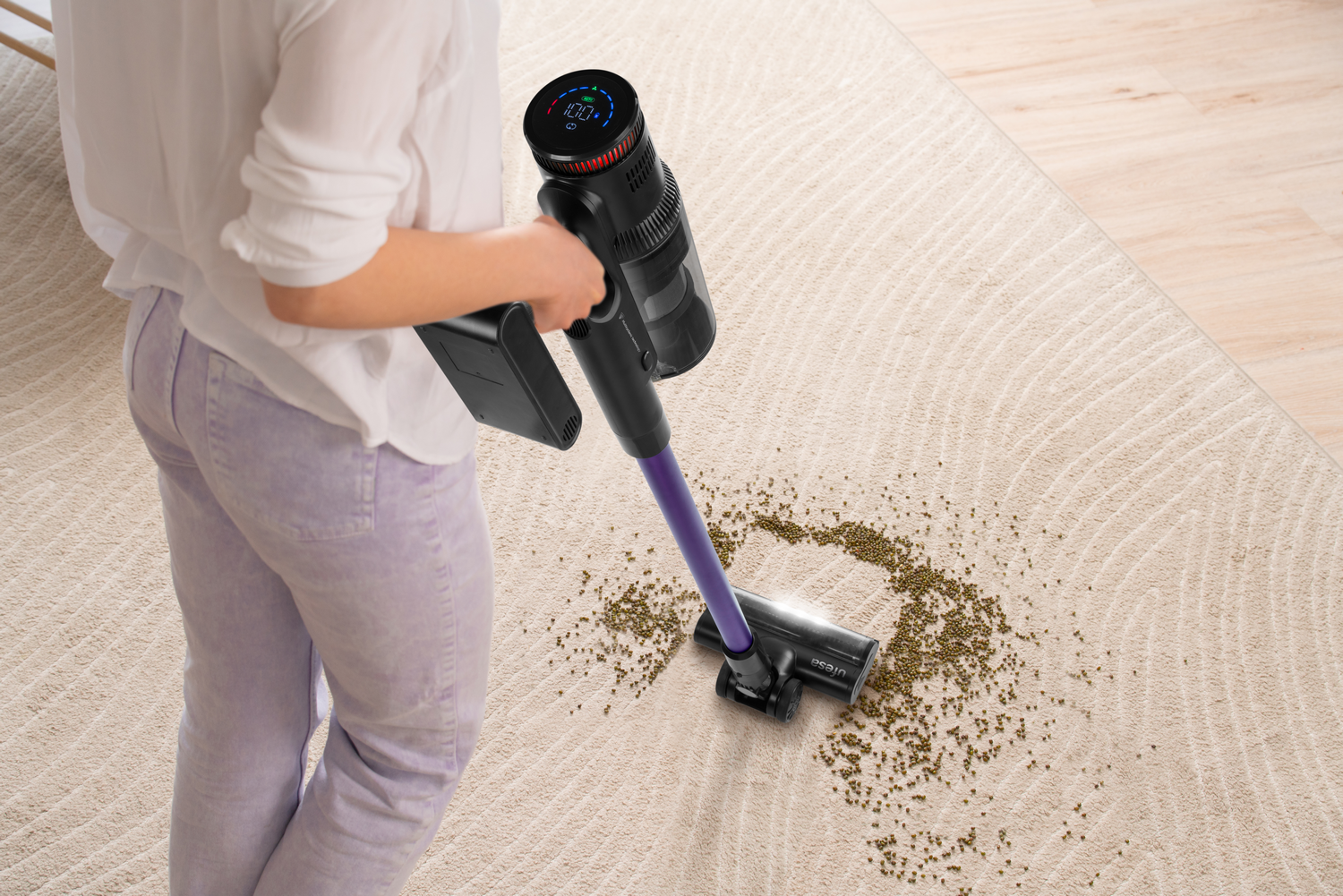 Una persona aspira una alfombra beige con una aspiradora inalámbrica. Hay suciedad en la alfombra.