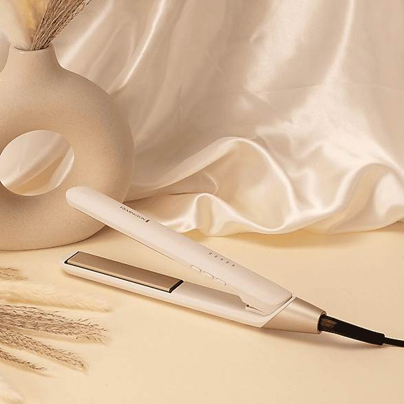 Una plancha para el cabello Remington blanca sobre un fondo claro con un jarrón y tela de seda.