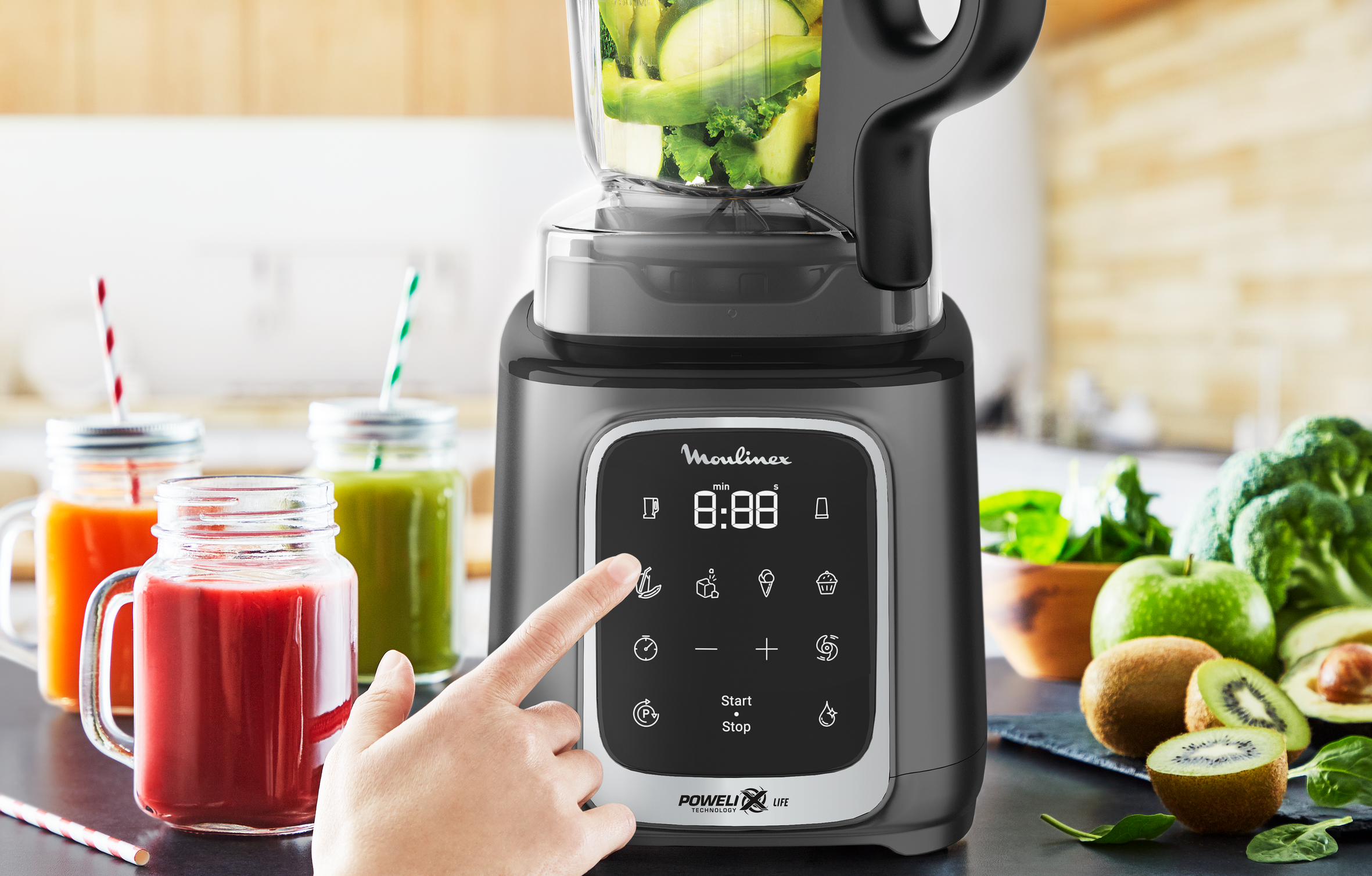 Licuadora con frutas y verduras dentro. Una mano toca la pantalla.