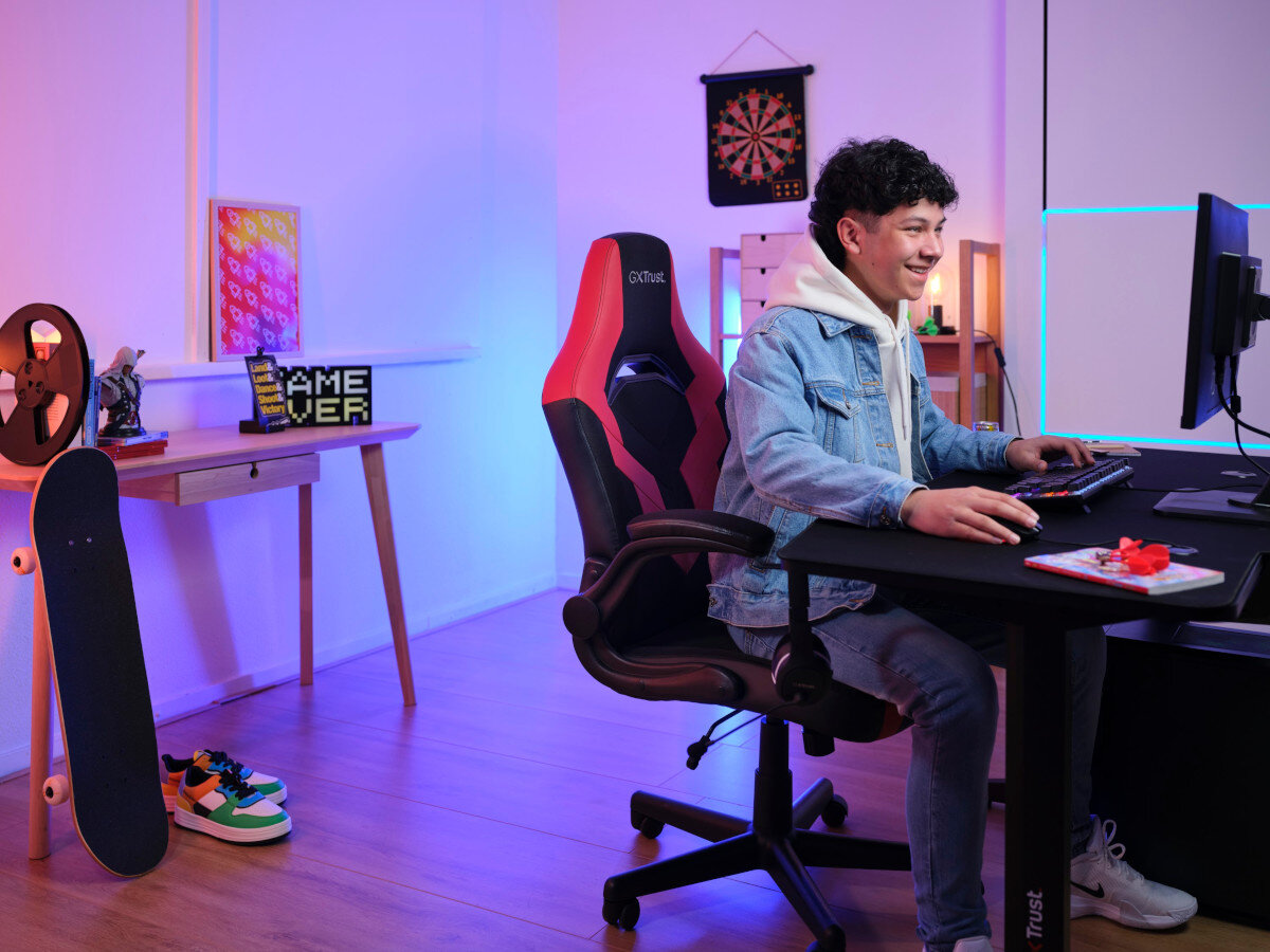 Nastolatek przy biurku, czerwone krzesło, monitor, deskorolka i przedmioty na biurku w kolorowym pokoju.