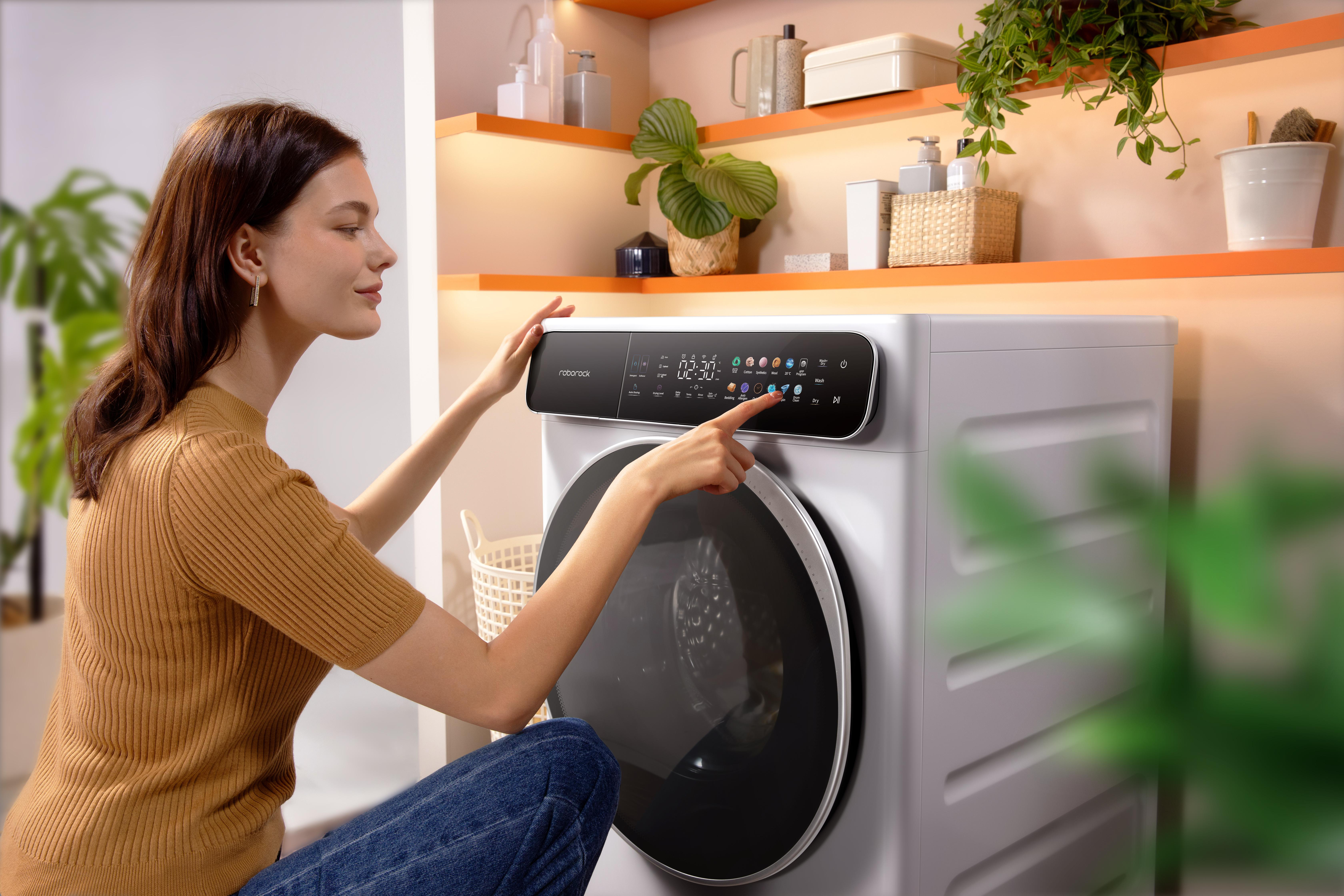 Eine Frau zeigt auf ein Bedienfeld einer Waschmaschine. Im Hintergrund sind Regale mit Pflanzen und Behältern zu sehen.