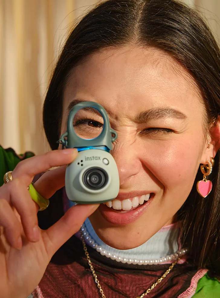 Joven guiñando un ojo mientras mira a través de una pequeña cámara instax. Sonriendo.