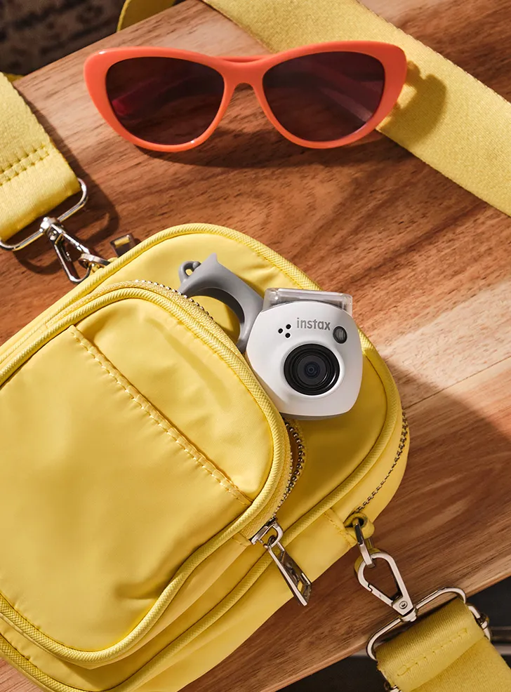 Bolso amarillo con gafas de sol y cámara Instax sobre superficie madera.