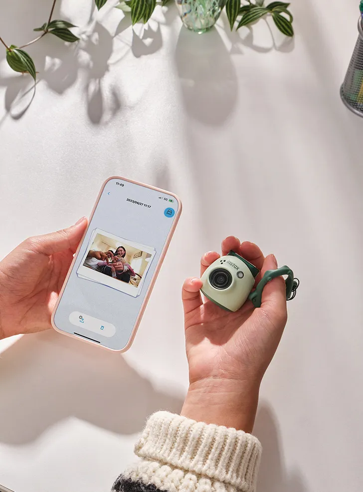 Manos sosteniendo un teléfono inteligente con una foto y una pequeña cámara verde. Superficie blanca.