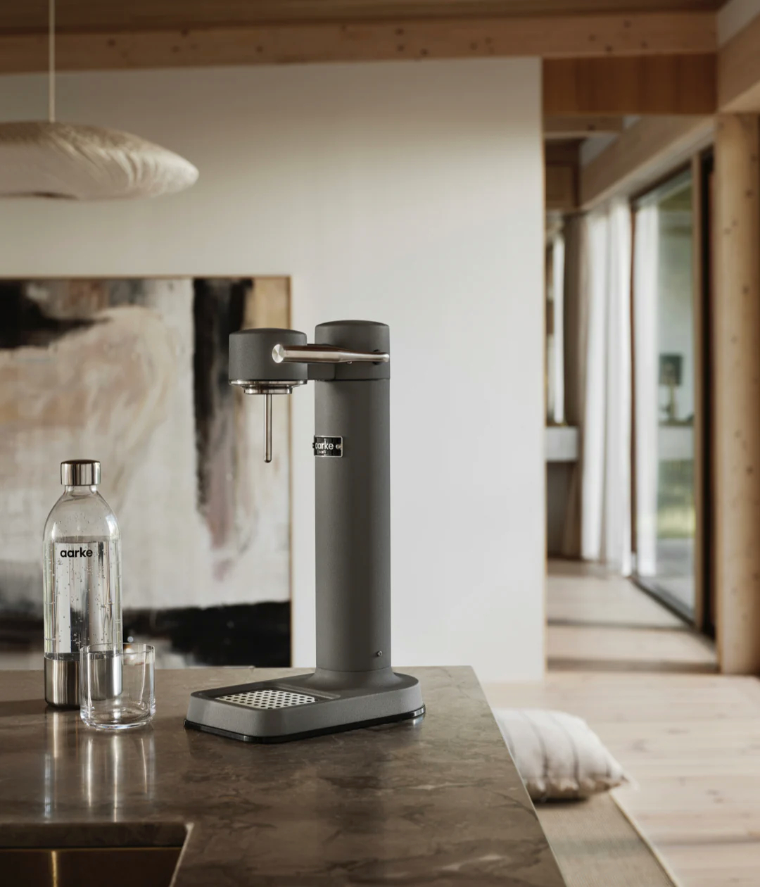 Ein grauer Wasserkohlesäureapparat neben einer Wasserflasche und einem Glas auf einer Steinarbeitsplatte. Modernes Interieur.