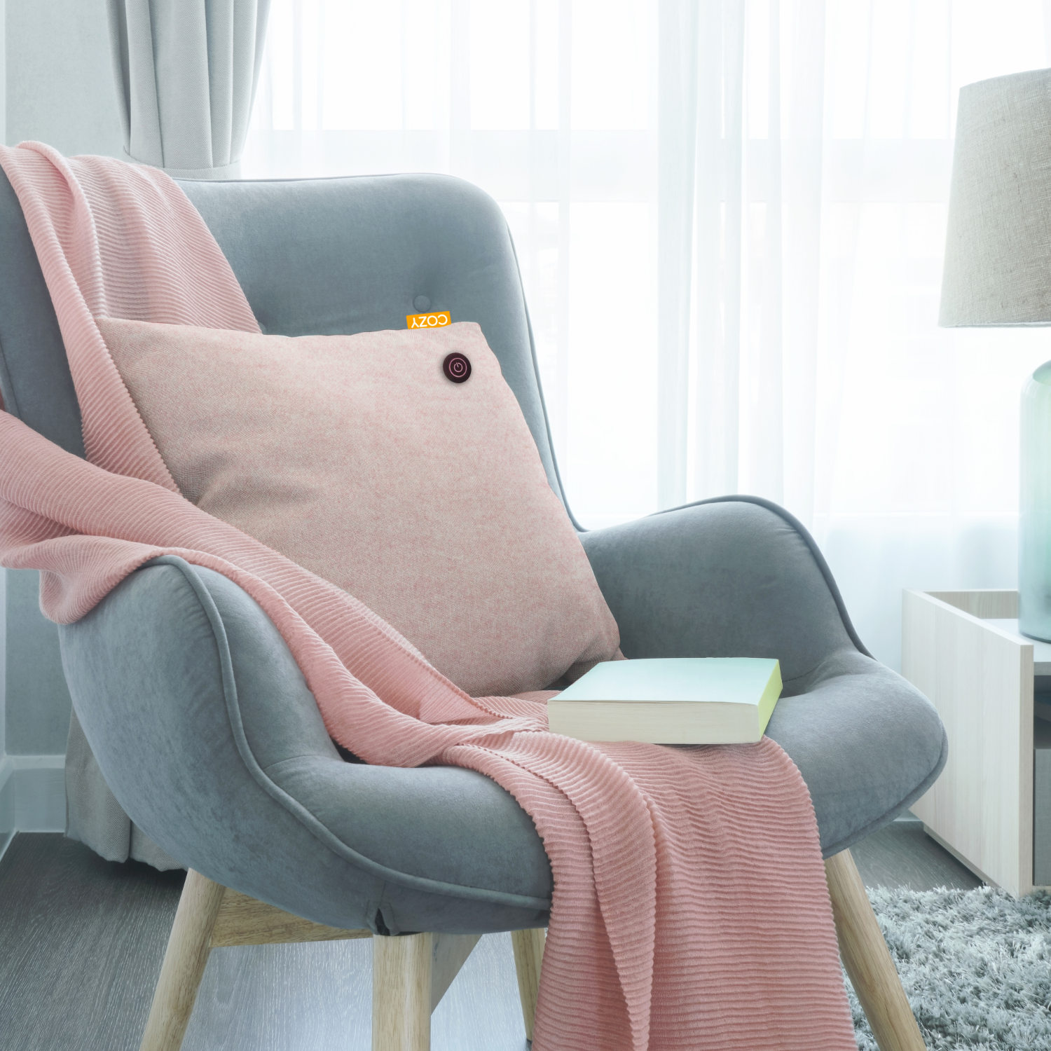 Grey armchair with a pink blanket and cushion, book and lamp.