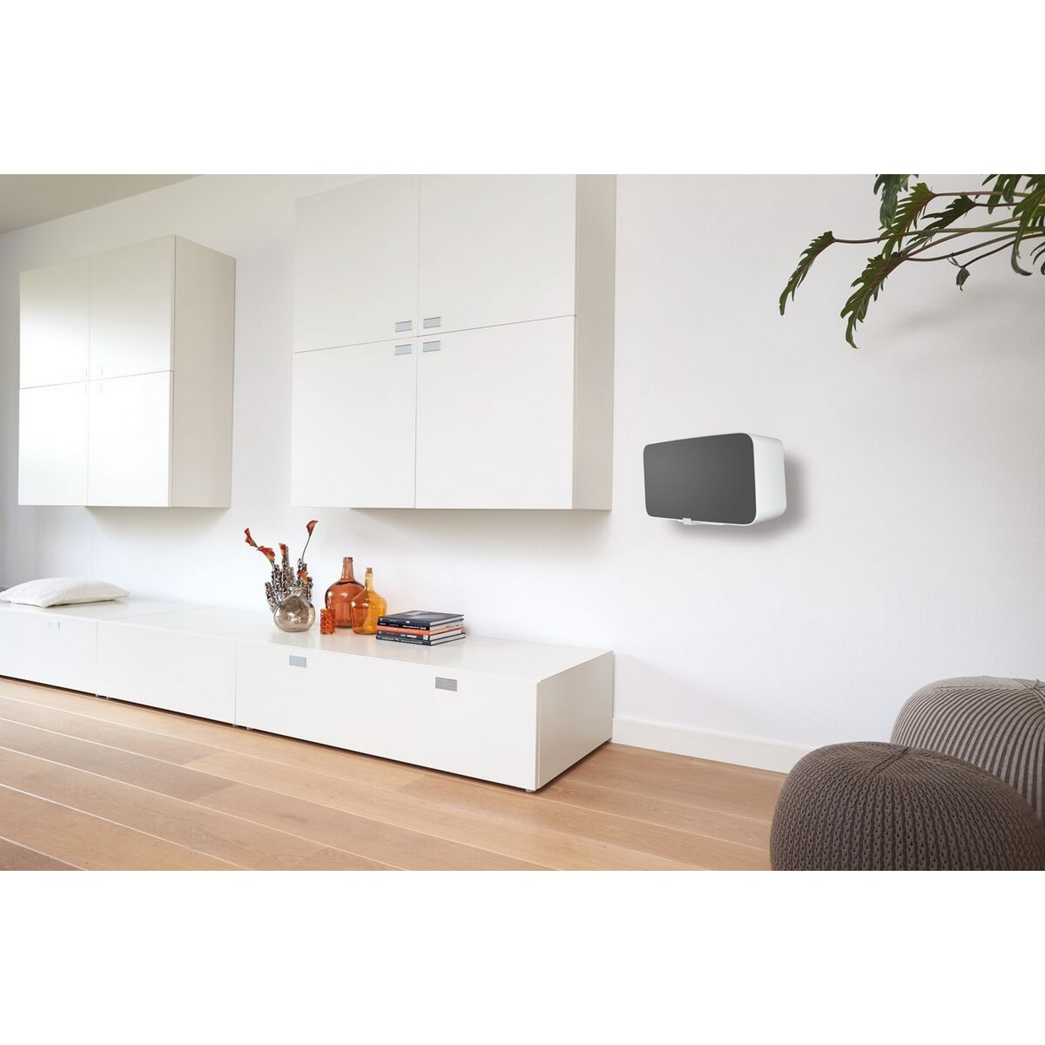 Mur blanc avec un haut-parleur noir et gris, des meubles blancs et un sol en bois. Une plante à droite.