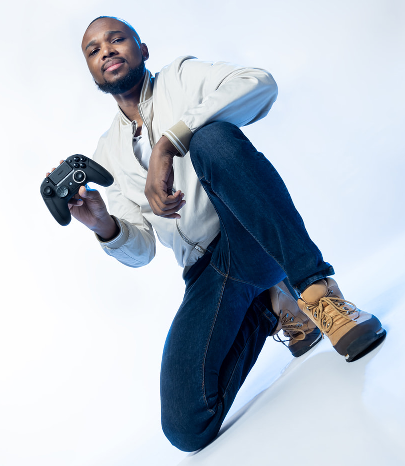 Mann kniet, hält einen schwarzen Controller, trägt Jeans, weiße Jacke und Stiefel.