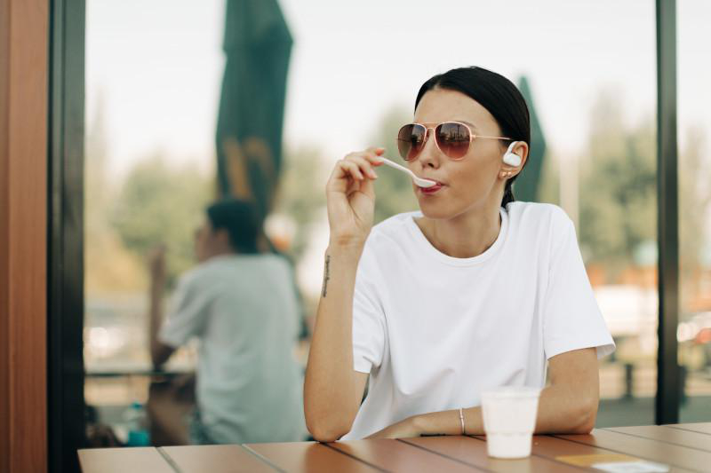 Eine Frau mit Sonnenbrille isst mit einem Löffel, sitzt an einem Tisch. Eine Tasse und verschwommener Hintergrund sind sichtbar.