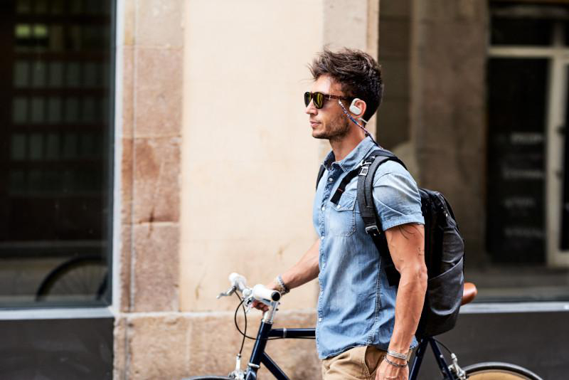 Ein Mann mit Sonnenbrille und Kopfhörern geht mit einem Fahrrad. Er trägt ein Jeanshemd und einen Rucksack.