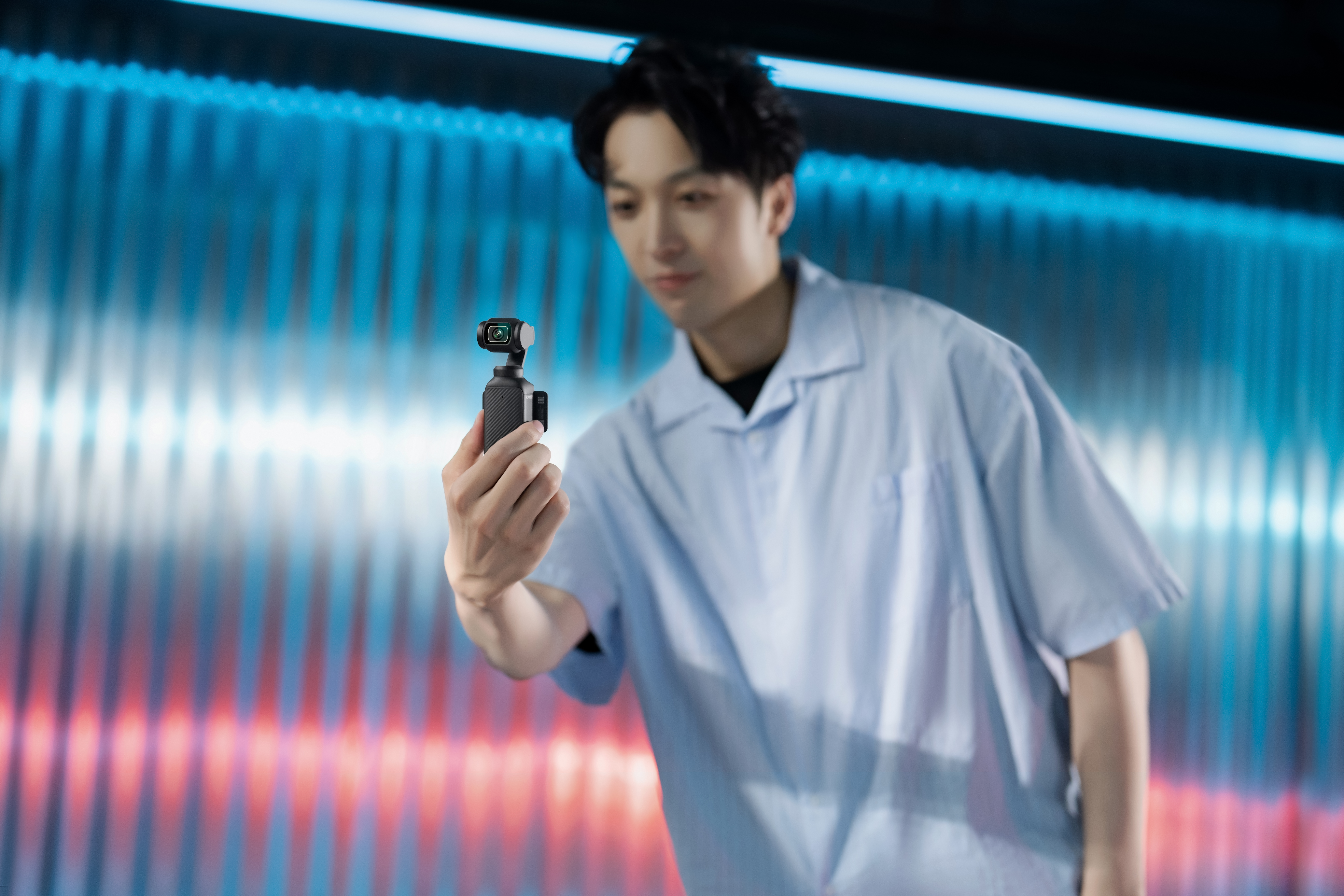 Un uomo tiene in mano una piccola fotocamera. Indossa una camicia blu. Lo sfondo è blu e rosso.