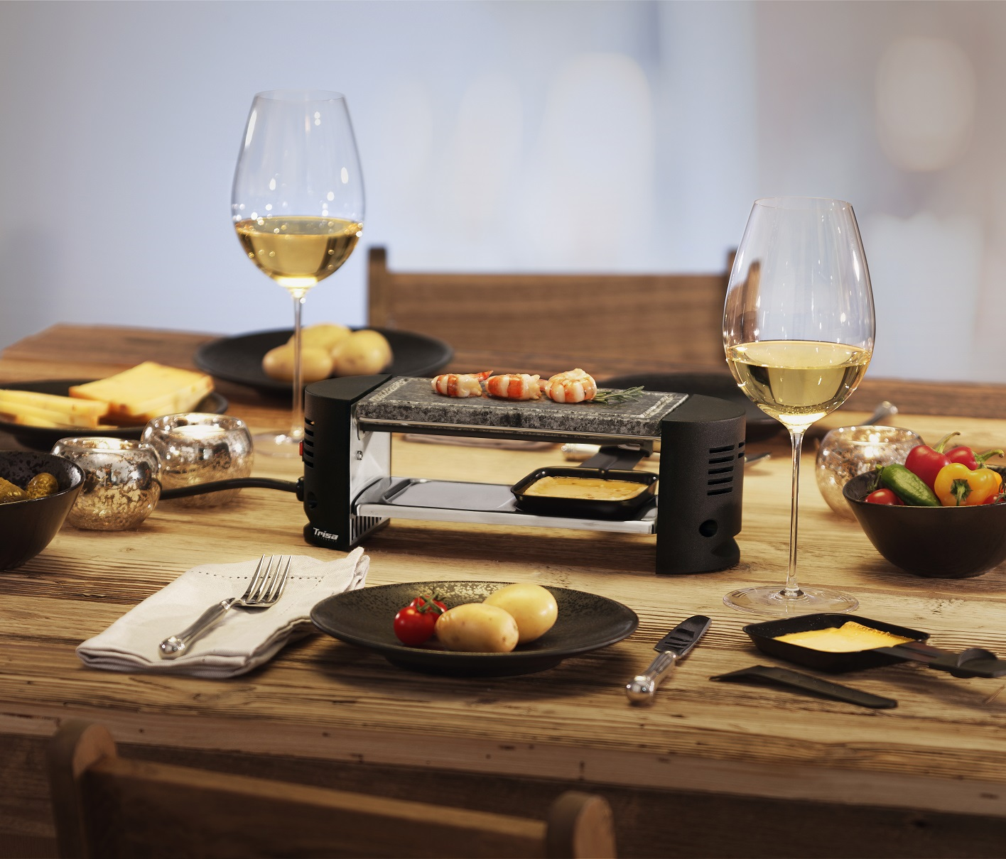 Tisch mit Raclette-Grill, Essen, Weingläsern und anderen Gegenständen.