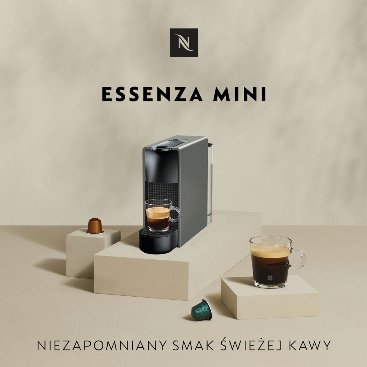 Ekspres do kawy Nespresso Essenza Mini. Szara maszyna z filiżankami i kapsułkami. Beżowe tło.