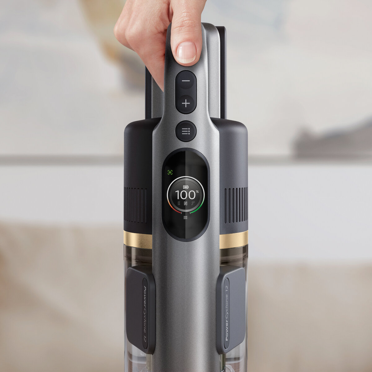 Ręka trzyma szary uchwyt odkurzacza. Wyświetlacz pokazuje baterię w 100%.