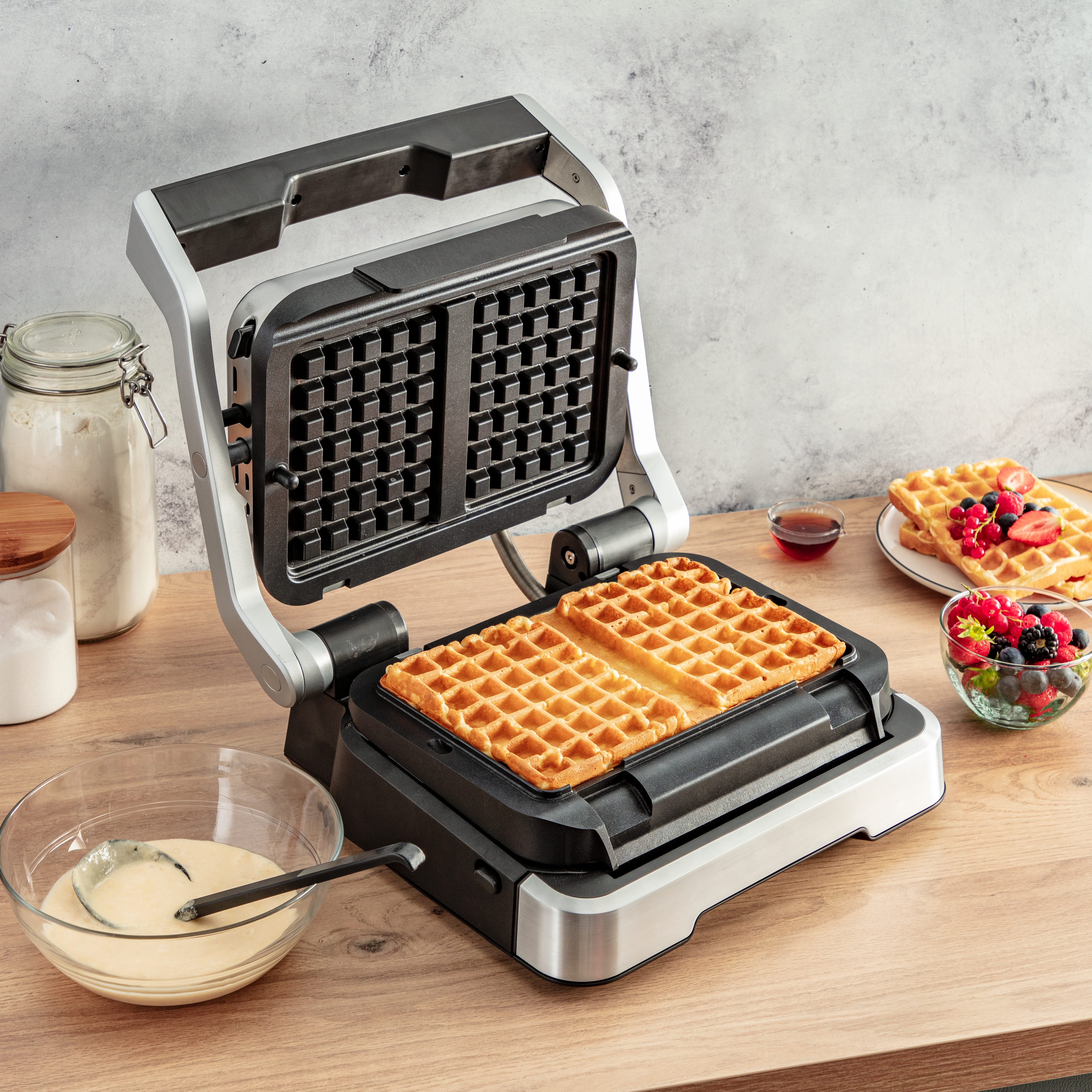 Ein Waffeleisen mit Waffeln, eine Schüssel mit Teig und Beläge auf einem Tisch.