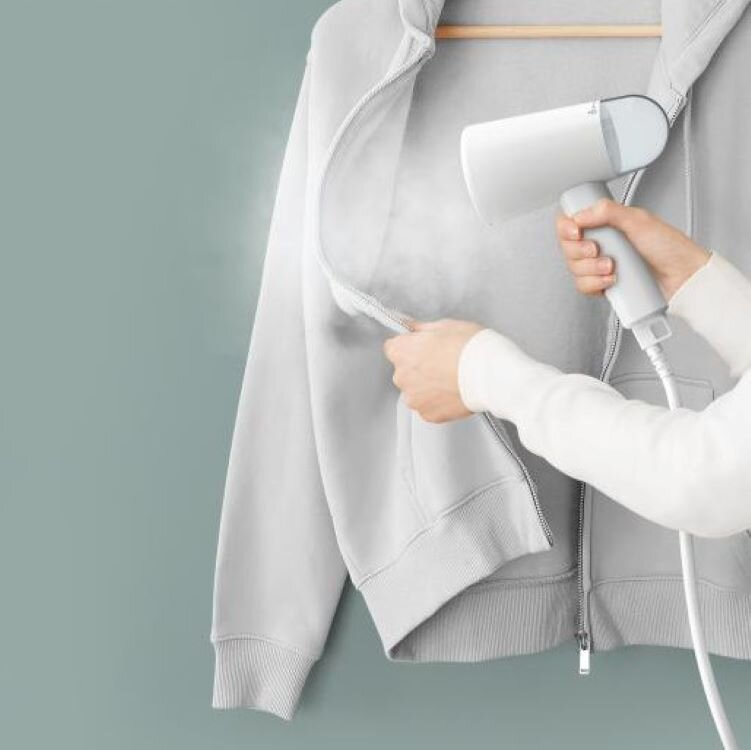 Osoba używająca parownicy do ubrań na szarej bluzie z kapturem, para unosi się w górę.