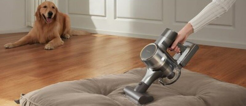 Osoba odkurzająca legowisko dla psa odkurzaczem ręcznym, a golden retriever obserwuje.