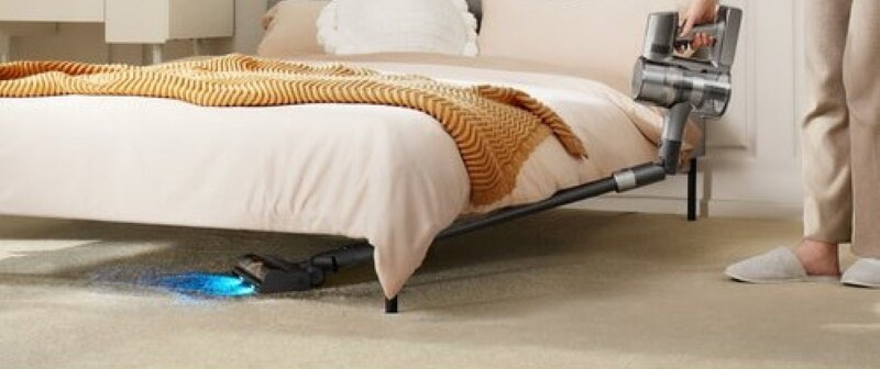 Osoba używająca odkurzacza pod łóżkiem. Odkurzacz świeci niebieskim światłem.