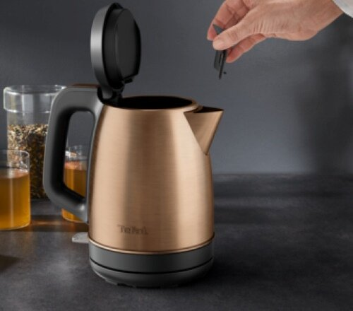 Miedziany czajnik. Czarna podstawa i uchwyt. Osoba zdejmująca pokrywkę. Tło blatu kuchennego.