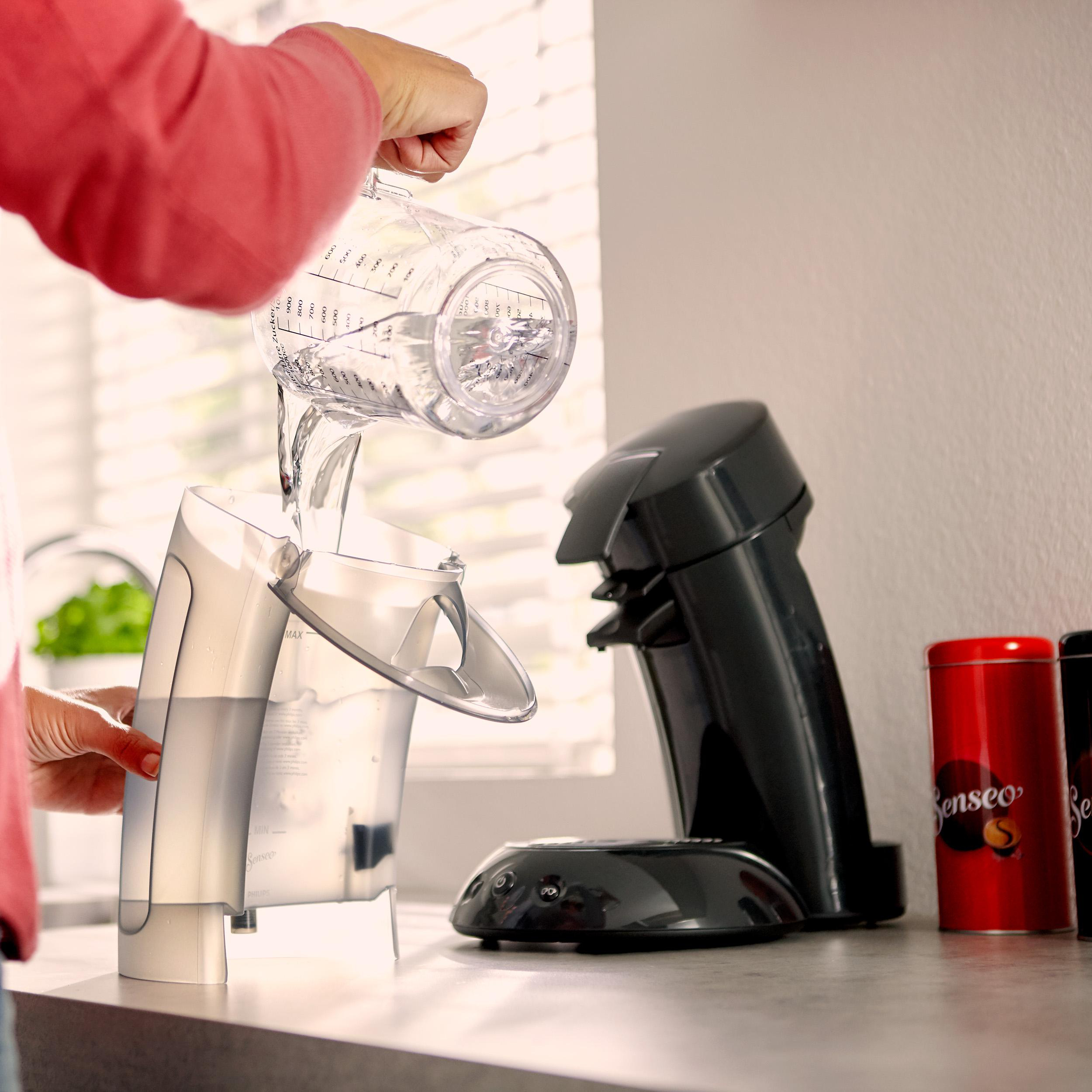 Eine Person gießt Wasser in eine schwarz-silberne Kaffeemaschine mit zwei roten Kaffeedosen.