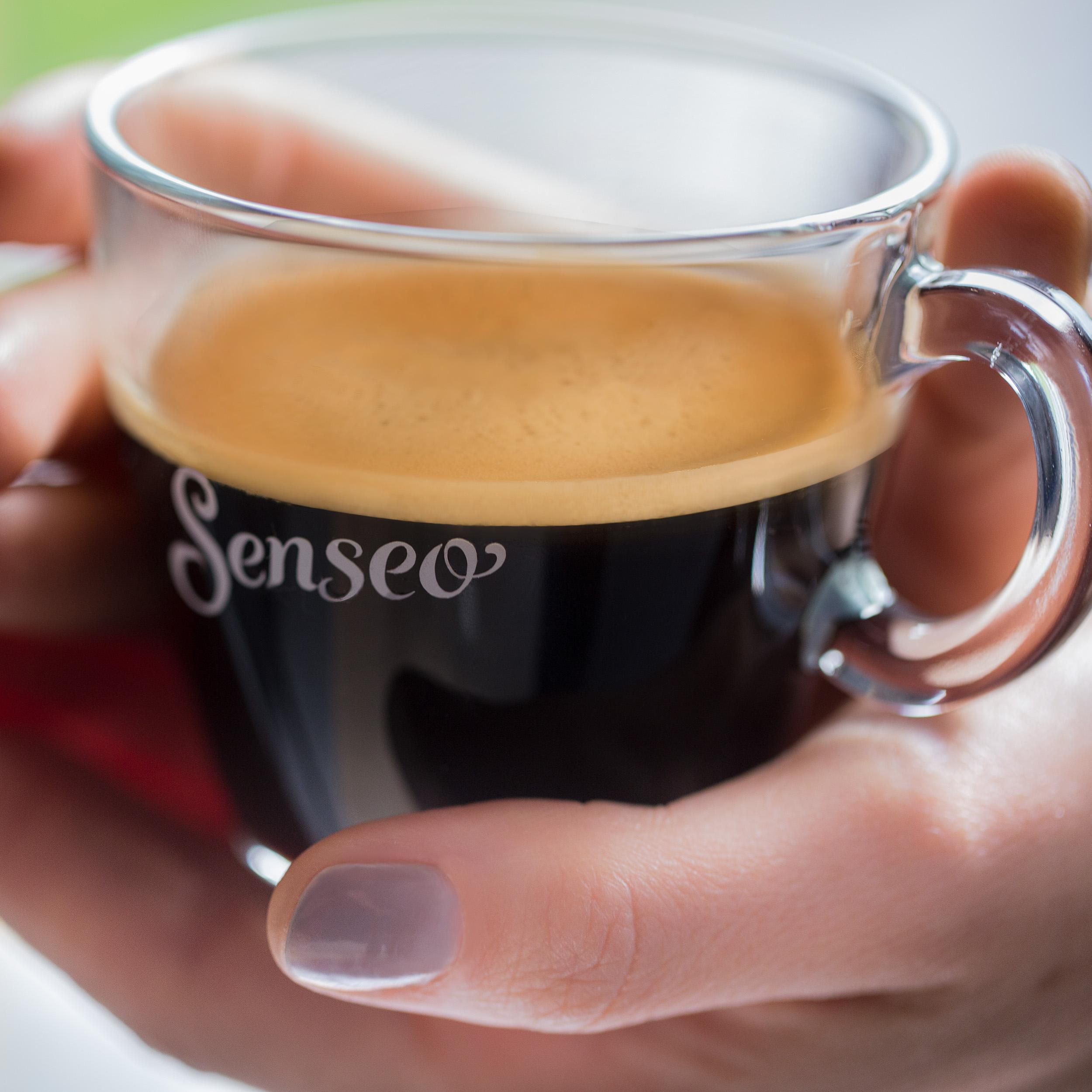 Ein Glas Kaffeetasse mit dem Wort „Senseo“ darauf, gehalten von Händen.