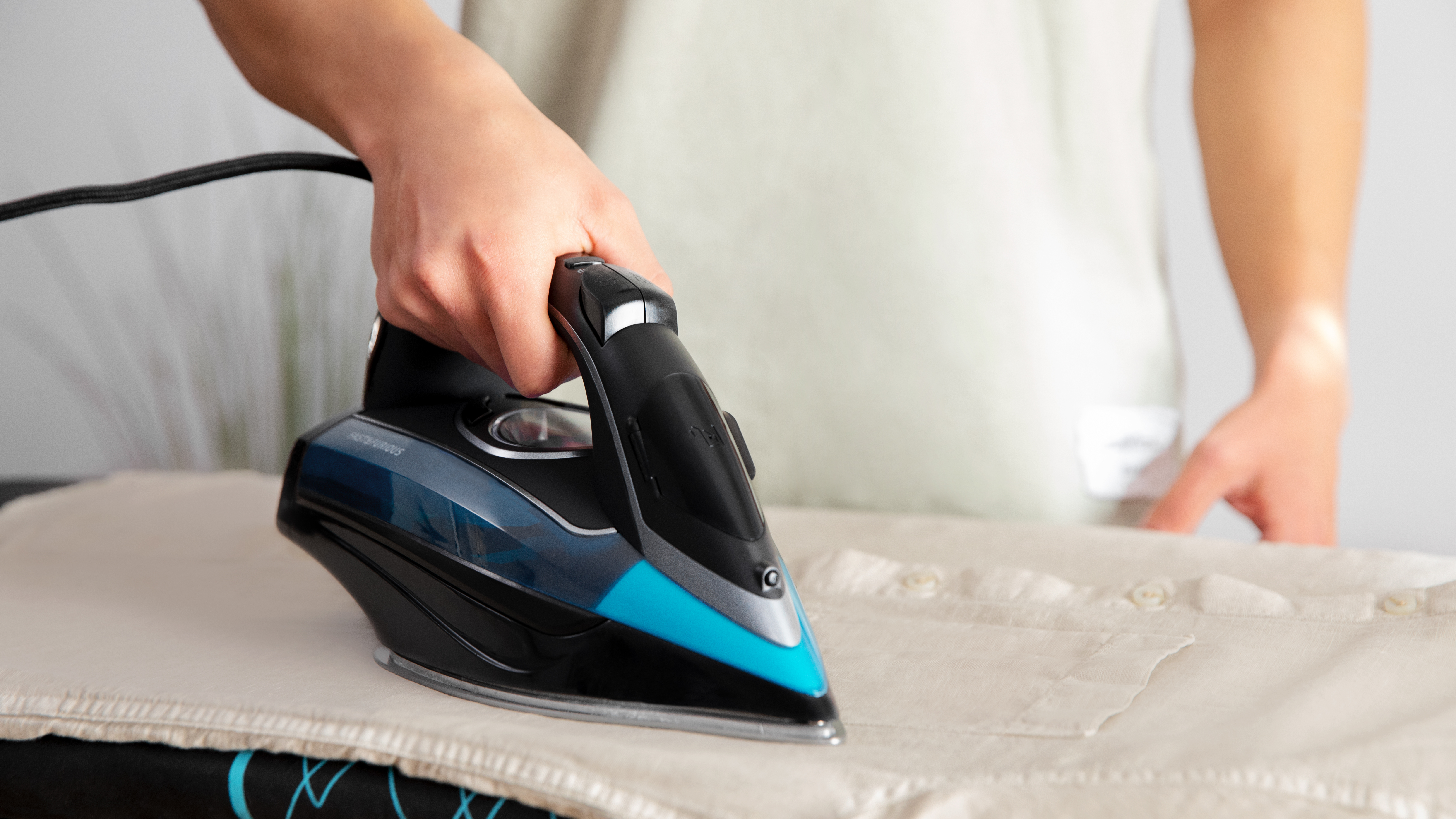 Una persona planchando una camisa blanca en una tabla de planchar; la plancha es azul y negra.
