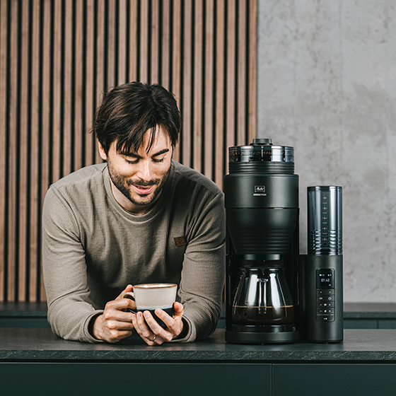 Mann hält eine Kaffeetasse neben einer Melitta-Kaffeemaschine und -Mühle auf einer Theke.