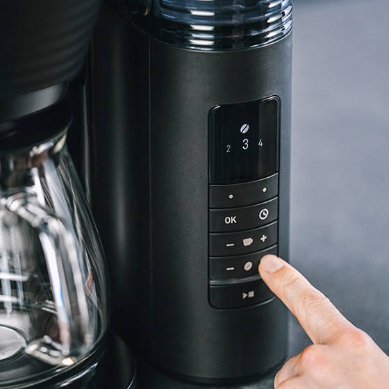 Schwarze Kaffeemaschine mit Bedienfeld, Finger drückt die Kaffeebohnen-Taste.