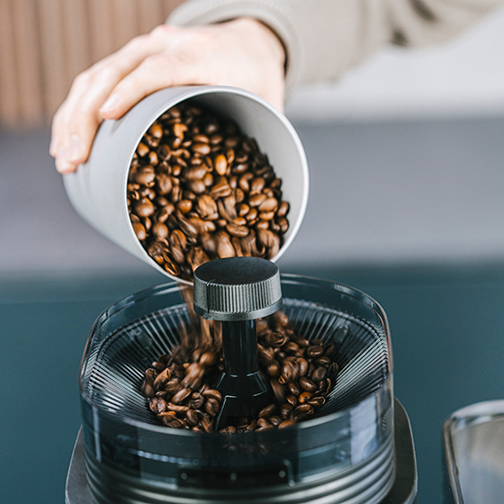Kaffeebohnen werden in eine Kaffeemühle gegossen. Die Mühle ist schwarz, die Bohnen braun.