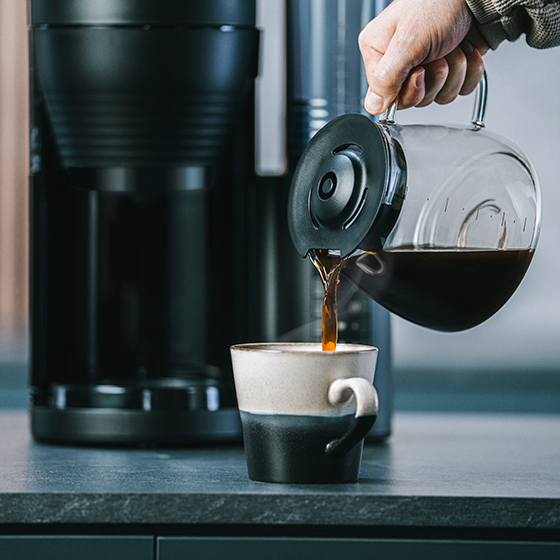 Kaffee wird aus einer Glaskaraffe in eine zweifarbige Tasse gegossen, in der Nähe einer Kaffeemaschine.