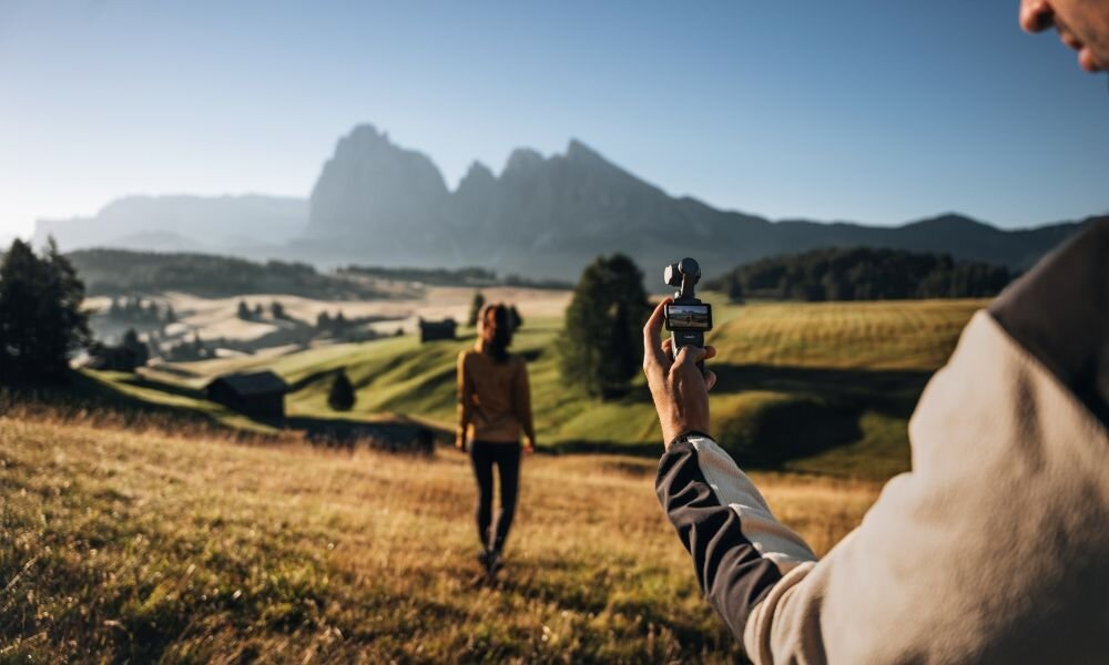Osoba filmująca kobietę idącą po polu, z górami w tle. Urządzenie filmowe uniesione.