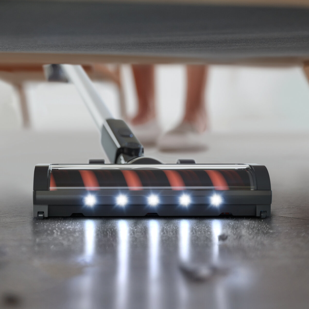 Głowica odkurzacza ze światłami, umieszczona na podłodze. Nogi i stopy rozmyte w tle.