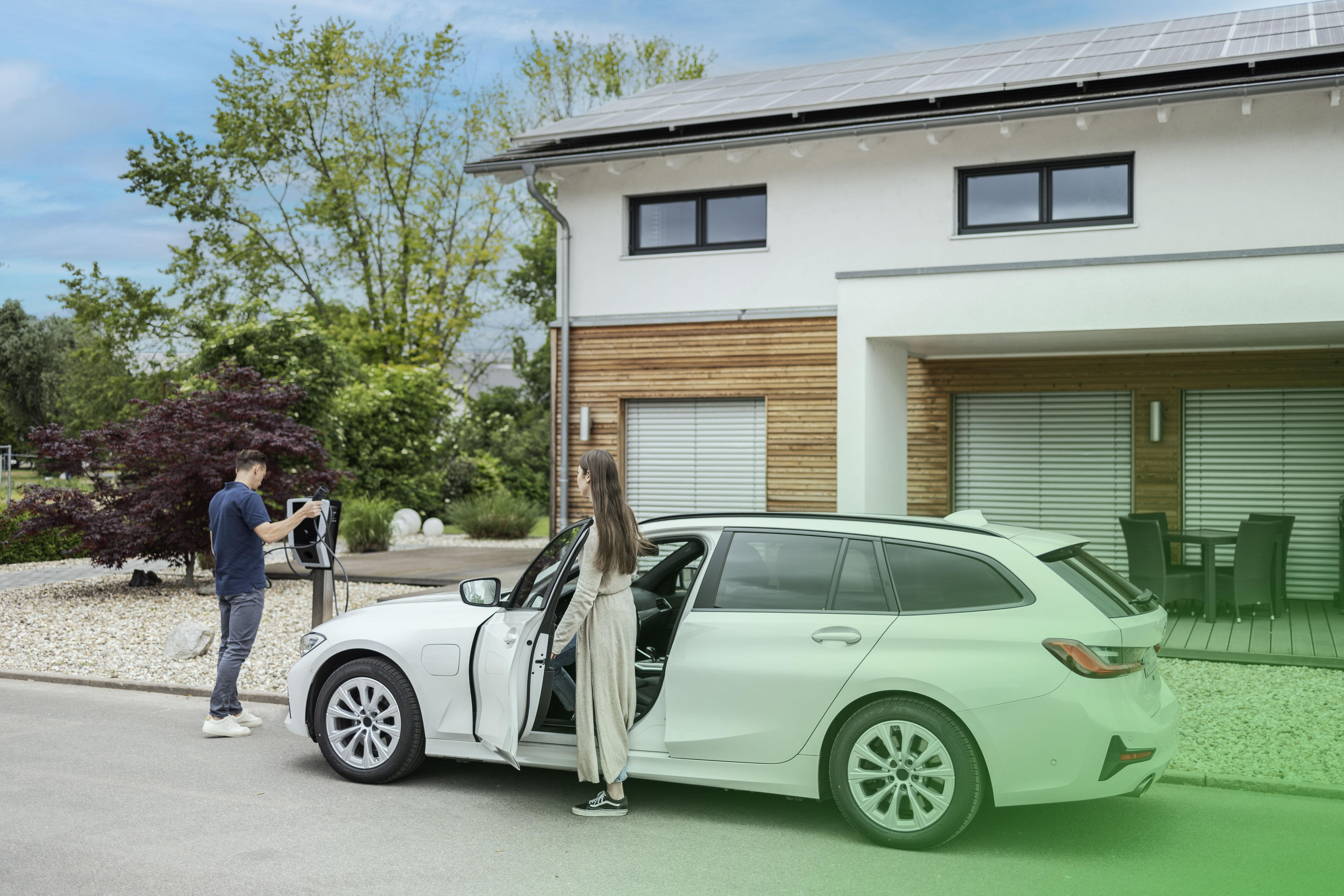 Ein Mann lädt ein Elektroauto auf, eine Frau steht daneben. Ein Haus und Sonnenkollektoren im Hintergrund.