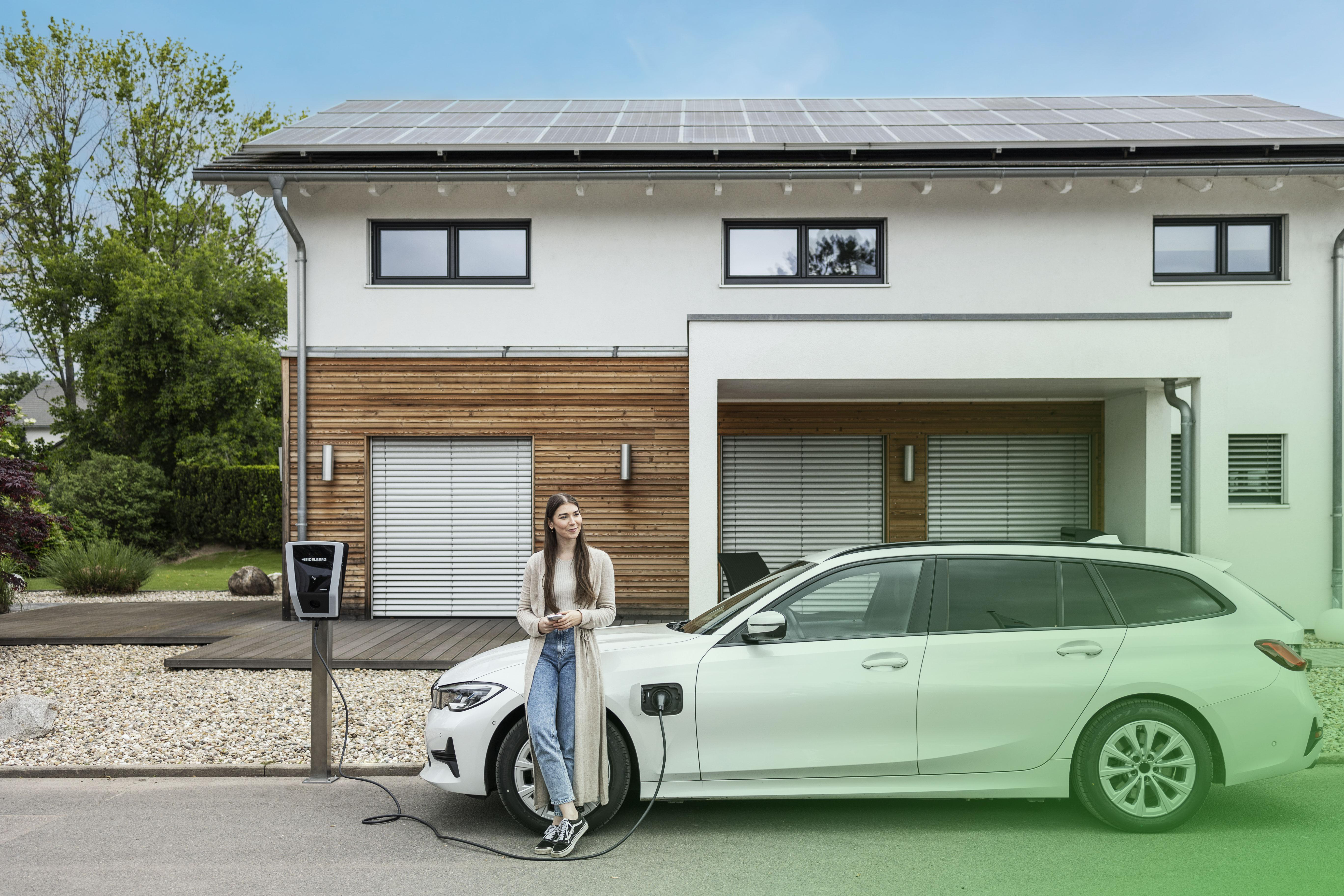 Frau lädt Elektroauto vor Haus mit Solaranlagen auf.