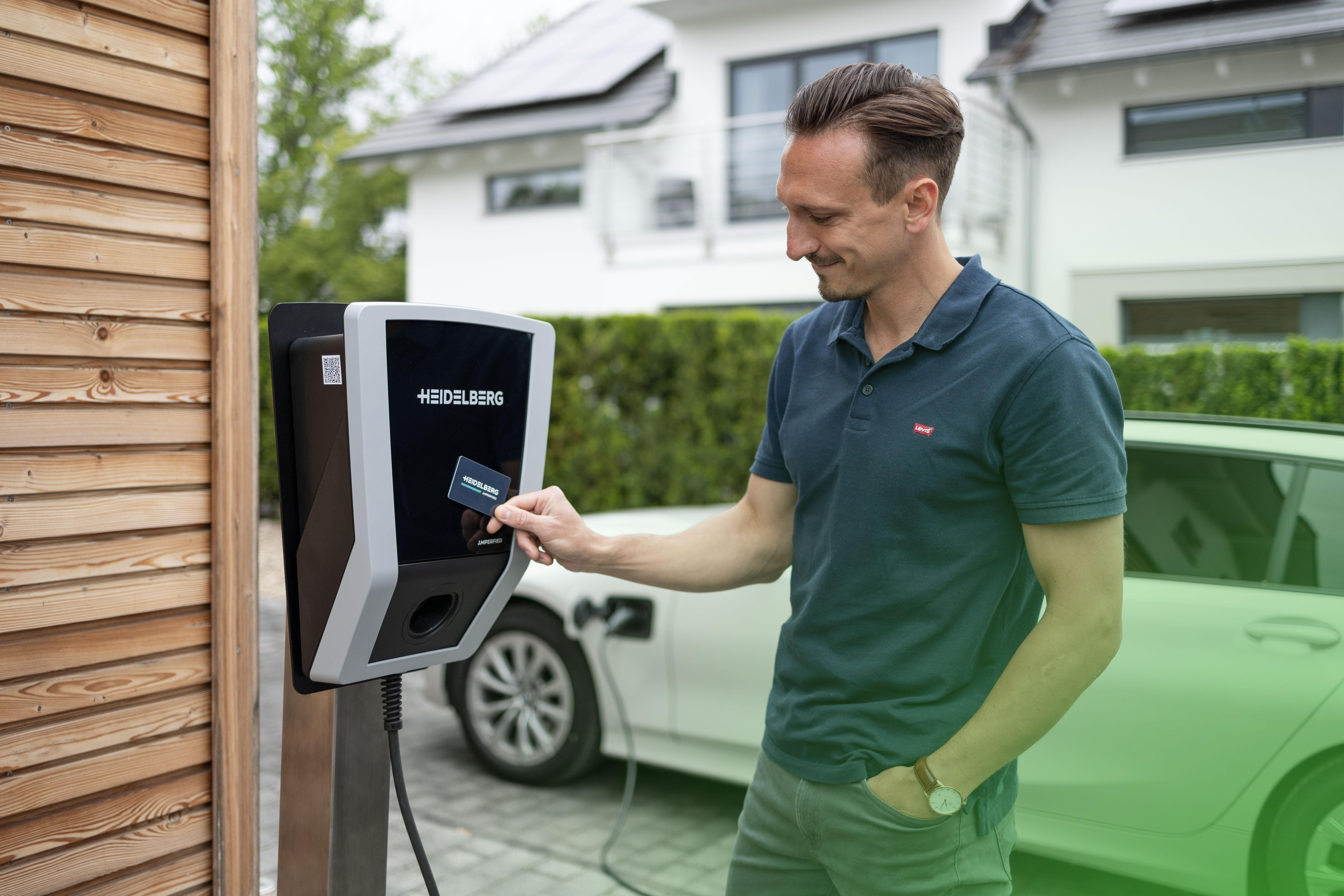 Mann benutzt Ladestation mit Karte, weißes Auto im Hintergrund.