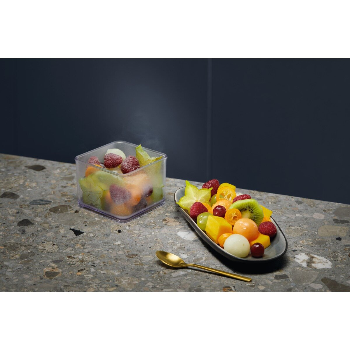 Kwadratowy pojemnik i talerz, wypełnione różnymi owocami, na kamiennej powierzchni.