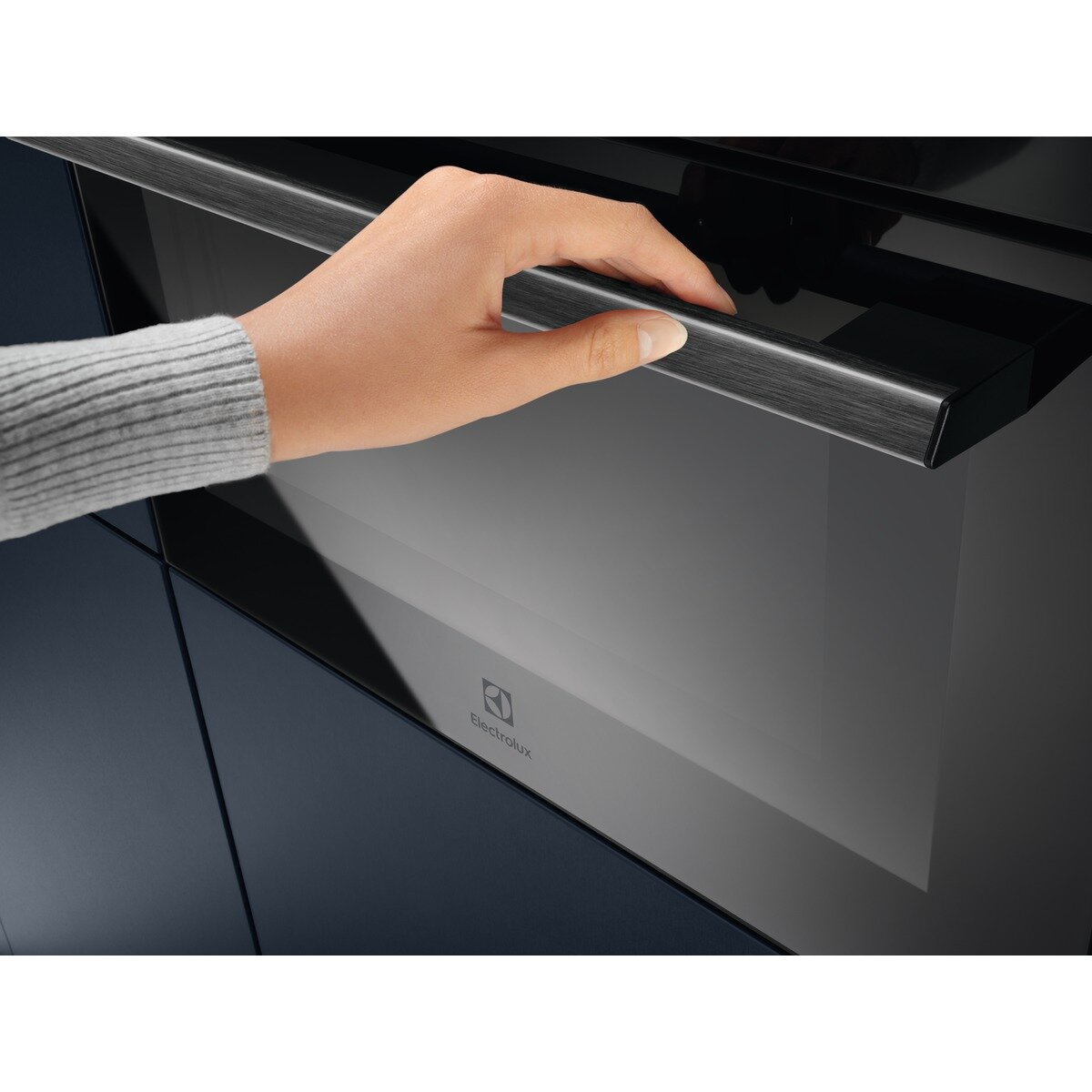 Dłoń otwiera czarne drzwi piekarnika ze srebrną klamką, widoczne logo Electrolux.