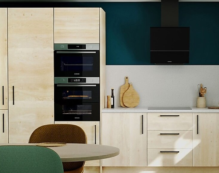 Nowoczesna kuchnia z jasnymi drewnianymi szafkami, stołem, piekarnikami i ciemnozieloną ścianą.