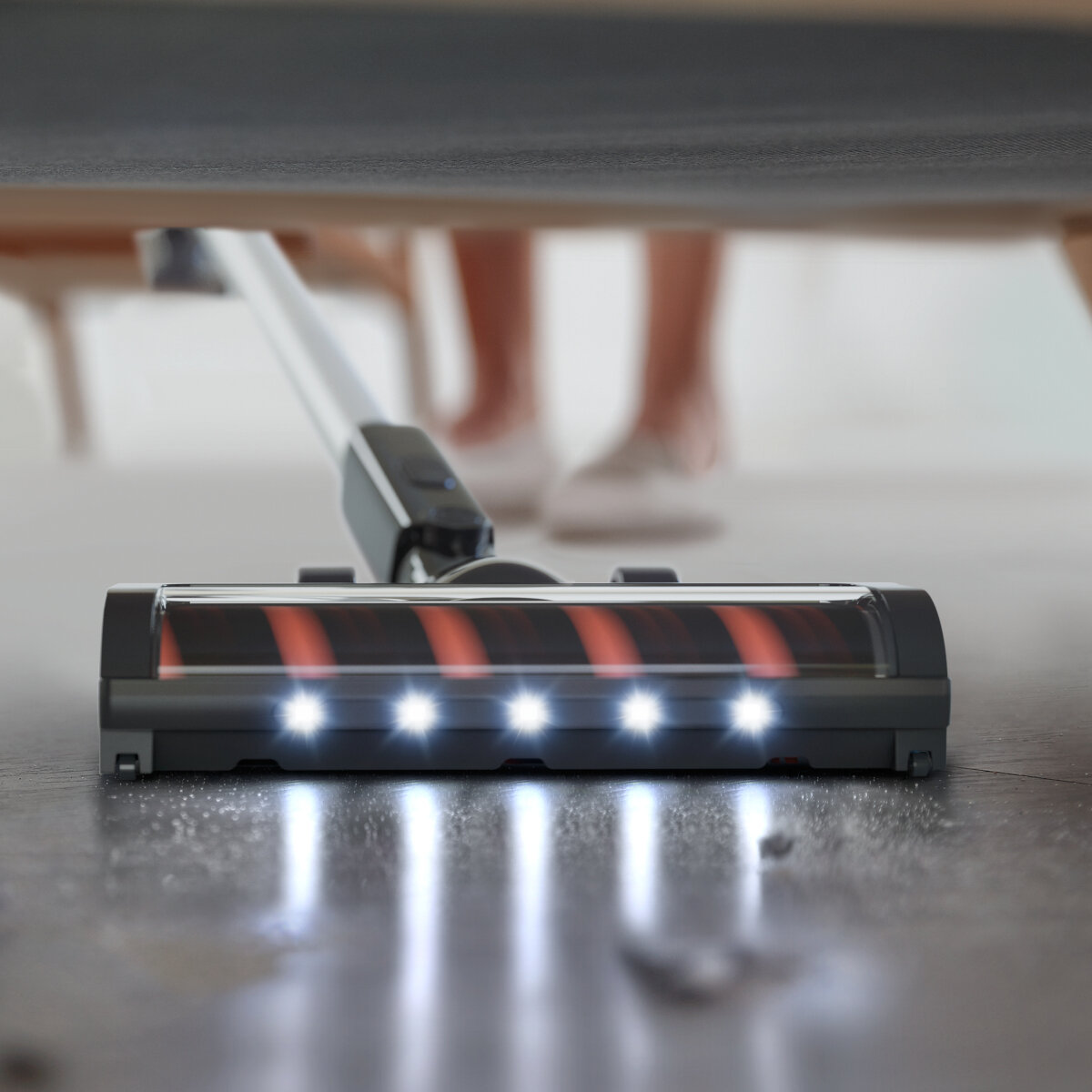 Głowica odkurzacza na podłodze. Światła włączone, osoba w tle. Szare i czerwone kolory.