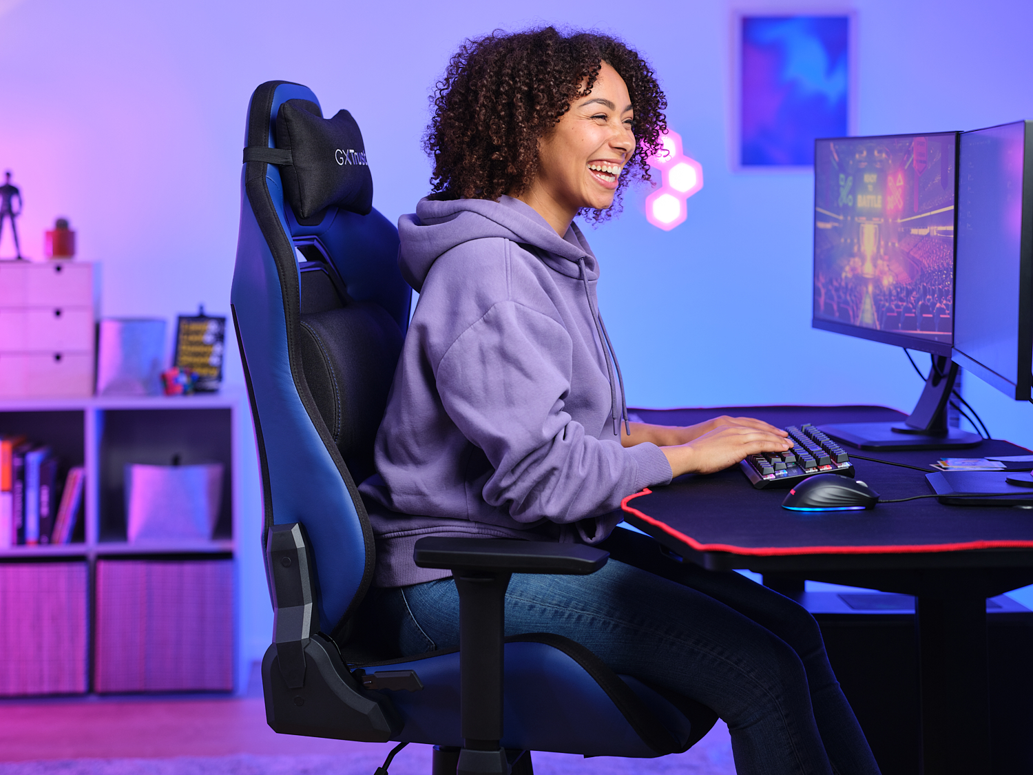 Une femme sourit en jouant. Elle est assise dans une chaise de jeu bleue, regardant un écran, avec un clavier devant elle.
