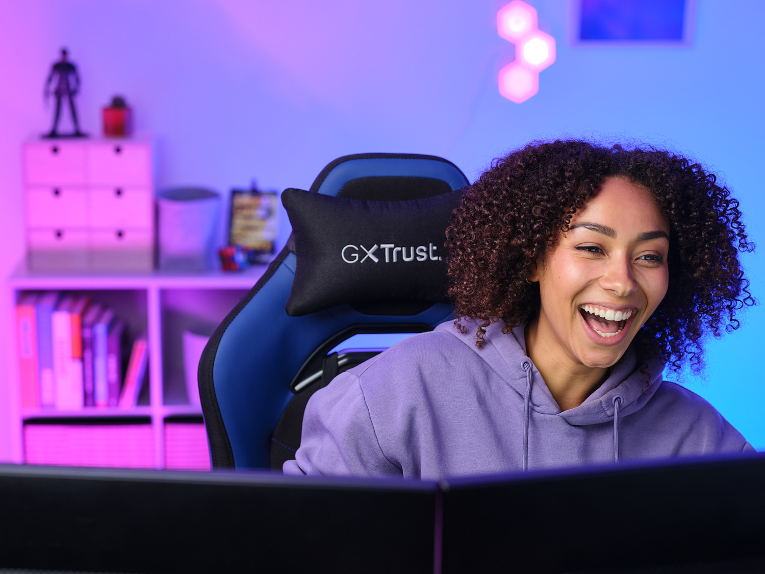 Femme souriant à un écran d'ordinateur. Elle est assise dans une chaise de jeu avec un design bleu et noir.