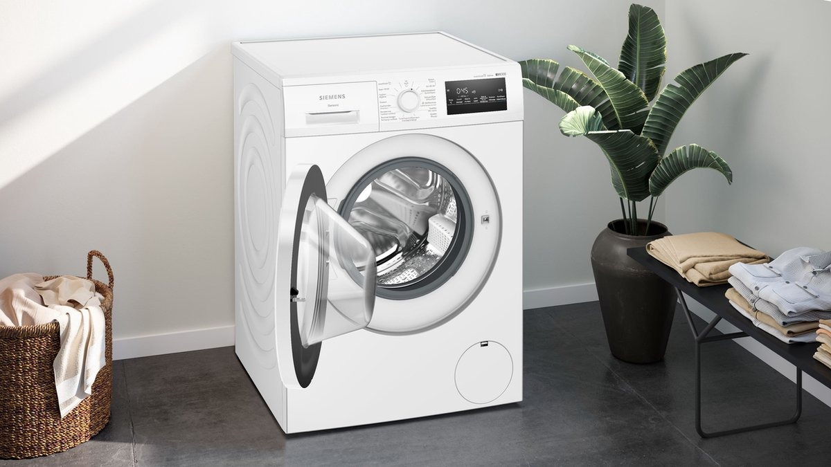 White Siemens washing machine with open door. Laundry basket and plant in the background.
