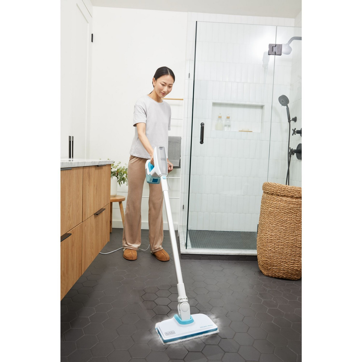 Een vrouw maakt een zwarte tegelvloer schoon met een stoommop in een badkamer.